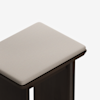 A close-up view of the corner of a wooden stool with a soft, light beige cushioned seat and dark brown legs against a white background.