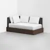A modern daybed with a dark wood base, charcoal gray upholstered backrest, white mattress, and four white pillows, placed in the corner of a minimalist room with white walls and a light gray floor.