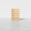 A minimalist wooden chest of five drawers stands on a light gray floor against a plain white background. The chest has a natural wood finish and no visible handles, giving it a modern, clean appearance.