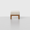 A modern ottoman with a light beige cushioned top and a simple wooden frame, placed on a smooth, light-colored floor against a plain white background.