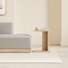 A minimalist wooden bed with white bedding is positioned next to a small, light wood nightstand against a plain white wall and light gray floor.