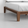 Close-up of a modern wooden bed frame with clean lines and tapered legs, supporting a mattress with white bedding, set on a light gray floor against a plain white wall.