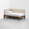 A modern daybed with a wooden frame, white mattress, white bedding, and a single pillow, positioned against two white walls in a minimalist room. The head and side backrests are upholstered in light beige fabric.