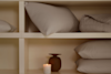 A neatly organized shelf holds folded bedding, white pillows, a beige comforter, and a lit white candle in a small vase, creating a calm and cozy atmosphere.