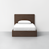 A simple, modern wooden bed frame with a tall headboard, made up with white bedding and a single white pillow, set against a plain white wall and gray floor.