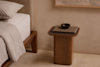 A minimalist wooden nightstand beside a bed holds a closed book and a small dark bowl. The bedding is neutral, and the wall and decor are in warm beige tones, creating a calm, modern atmosphere.