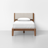 A single wooden bed with a beige upholstered headboard, white bedding, and a single pillow, set against a plain white wall in a minimalistic room.
