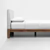 A minimalist bed with a light gray upholstered headboard, white bedding, and a wooden frame, positioned against a plain white wall on a smooth white floor.
