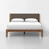 A modern wooden bed frame with a dark upholstered headboard, white bedding, and two white pillows, set against a plain white wall and light gray floor.