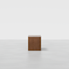 A single, square wooden cube sits on a smooth, light gray floor against a plain white background. The cube has a simple, minimalist design with no visible decorations.