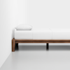 A minimalist wooden bed frame with white bedding and a single white pillow, positioned against a white wall on a light, smooth floor.