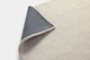 A close-up of a light beige textured rug with one corner folded over, revealing a dark gray underside. The rug is set against a plain white background.