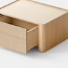 A minimalist, light wood nightstand with a single drawer partially open, set against a plain white background and light gray floor.