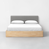 A neatly made bed with white bedding, two pillows, a light wooden frame, and a gray upholstered headboard against a white wall in a minimalist room.