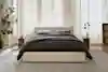 A modern bedroom with a beige upholstered bed, dark bedding, two pillows, and two wooden nightstands. The room has large windows with sheer white curtains, a tall plant, dried flowers, and a light gray rug.