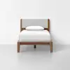 A single wooden bed with a light gray upholstered headboard, neatly made with white bedding and a single white pillow, set against a plain white wall and floor.