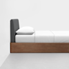 A minimalist bed with a dark gray upholstered headboard, white bedding, and a wooden base is placed against a white wall on a light gray floor.