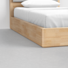 A close-up of a modern wooden bed frame with white bedding and pillow, positioned on a smooth, light gray floor.