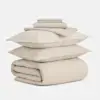 A neatly stacked bedding set includes two folded pillowcases, a flat sheet, a fitted sheet, two pillows, and a rolled-up comforter, all in a light beige color.