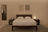A neatly made bed with white bedding and two pillows sits between two dark wood nightstands, each with a lamp. Minimalist decor includes a mirror, dresser, and two abstract artworks on the beige wall.
