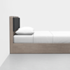 A minimalist wooden bed with a light wood frame, black cushioned headboard, white pillow, and neatly made white bedding, positioned against a plain white wall on a gray floor.