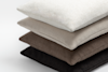 A neatly stacked pile of four textured fabric cushions in neutral colors: white, beige, brown, and dark brown, arranged from top to bottom on a plain white background.