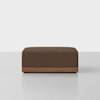 A modern, square ottoman with dark brown upholstery and a wooden base, placed on a light gray floor against a plain white background.