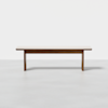 A simple wooden bench with a light-colored top and darker legs, placed on a smooth, light gray floor against a plain white background.