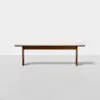 A simple wooden bench with a light-colored top and darker legs, placed on a smooth, light gray floor against a plain white background.