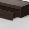 A close-up of a dark wood bed frame with a large, open storage drawer underneath, set on a light-colored floor.