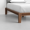 Close-up of a wooden bed frame with square legs and a white mattress, shown in a minimal, modern room with a light gray floor and a light background.
