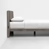 A minimalist wooden bed frame with a cushioned fabric headboard, neatly made with white bedding and pillow, is placed against a plain white wall in a bright, modern room.