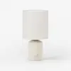 A modern table lamp with a cylindrical white fabric shade and a simple, matte cream base featuring a small round button on the front, set against a plain white background.