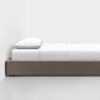 A neatly made bed with a gray upholstered frame, white sheets, and a single white pillow, positioned against a plain white wall on a light-colored floor.