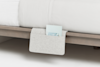A light gray felt bedside pocket hangs from a wooden bed frame, partially filled with a book. The white bedding and minimalist design create a clean, modern look.