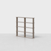 A minimalist wooden bookshelf with three columns and four rows, creating twelve open compartments, standing on a light grey floor against a plain white background.
