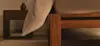 Close-up of a wooden bed frame with beige bedding draped over the side, next to a nightstand holding a vase and a book, in a softly lit bedroom.