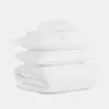 A neatly stacked set of all-white bedding, including two pillows, a folded duvet, and folded sheets, against a plain white background.
