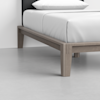A close-up of a modern wooden bed frame with light wood finish, white bedding, and a black headboard, set on a light gray floor in a minimalistic room.