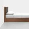 Side view of a modern wooden bed with a dark cushioned headboard, white pillow, and neatly made white bedding, positioned against a white wall on a gray floor.