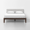 A modern bed with a light gray upholstered headboard, two white pillows, and a white bedspread on a dark wooden frame, set against a plain white wall and floor.