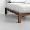 A close-up view of a bed with a wooden frame and white bedding, showing the corner of the bed and light gray flooring.