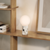 A modern table lamp with a round frosted glass shade and a marble base sits on a wooden shelf next to stacked books and a framed artwork on a neutral wall.
