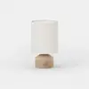 A small, modern table lamp with a beige cylindrical fabric shade and a round, matte beige base featuring a single button on the front, set against a plain white background.