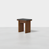A small wooden stool with a rectangular black cushioned seat and wide, sturdy legs, placed on a plain light-colored floor against a white background.