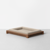 A rectangular pet bed with a wooden frame and beige cushioned center sits on a light gray floor against a plain white background.