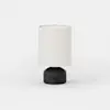 A modern table lamp with a cylindrical white fabric shade and a round, matte black base featuring a small circular button on the front, set against a plain white background.