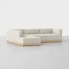 A modern, L-shaped sectional sofa with light beige upholstery and a wooden base, set against a plain, light gray background.