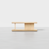 A minimalist wooden coffee table with a rectangular top, an off-center vertical panel, and an open shelf beneath, set against a plain white background.