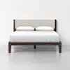 A modern bed with a dark wooden frame, beige upholstered headboard, white bedding, and two white pillows, set against a plain white wall and floor.